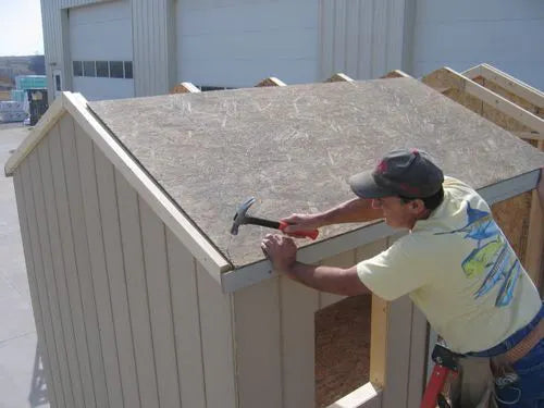 a man using a hammer to nail down the roof panel of a little cottage co storage shed kit