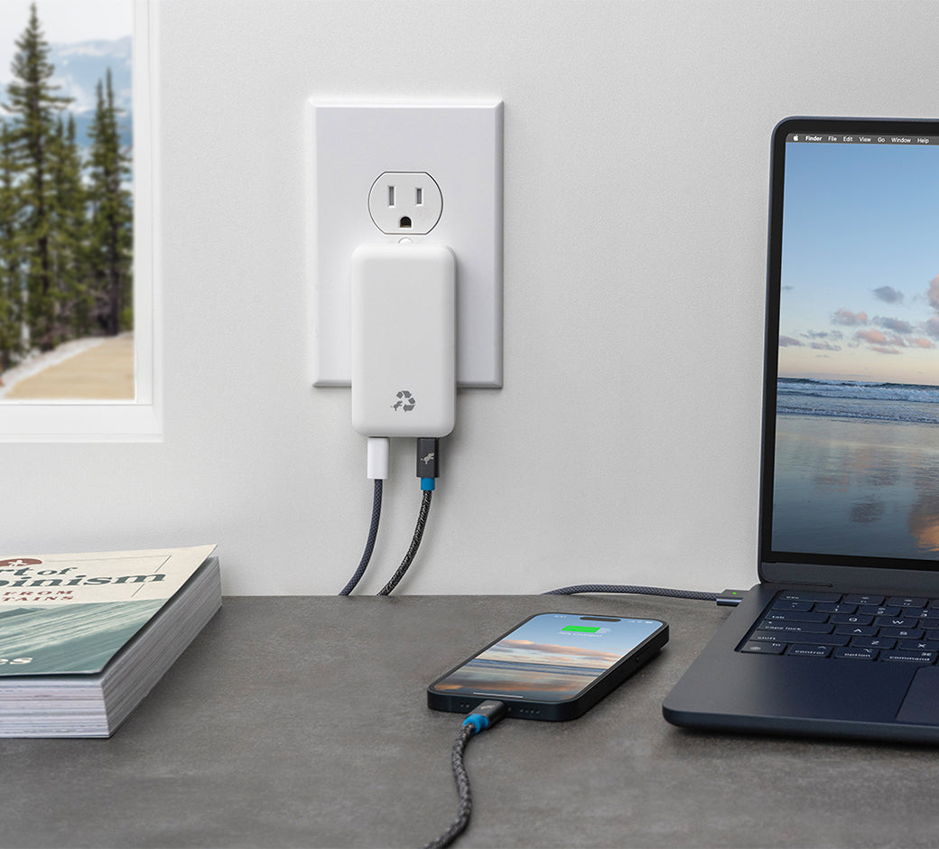 White dual-port wall charger powering a phone and laptop on a desk near a window.