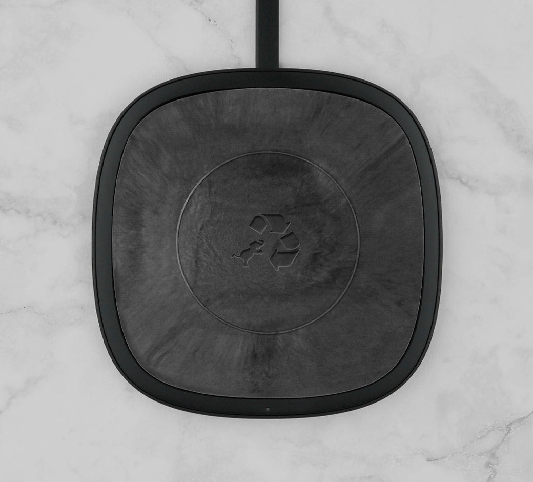 A black wireless charging pad with a subtle recycling logo in the center, placed on a marble surface.