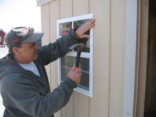 A man is installing a window on the siding of a Little Cottage Co. shed.