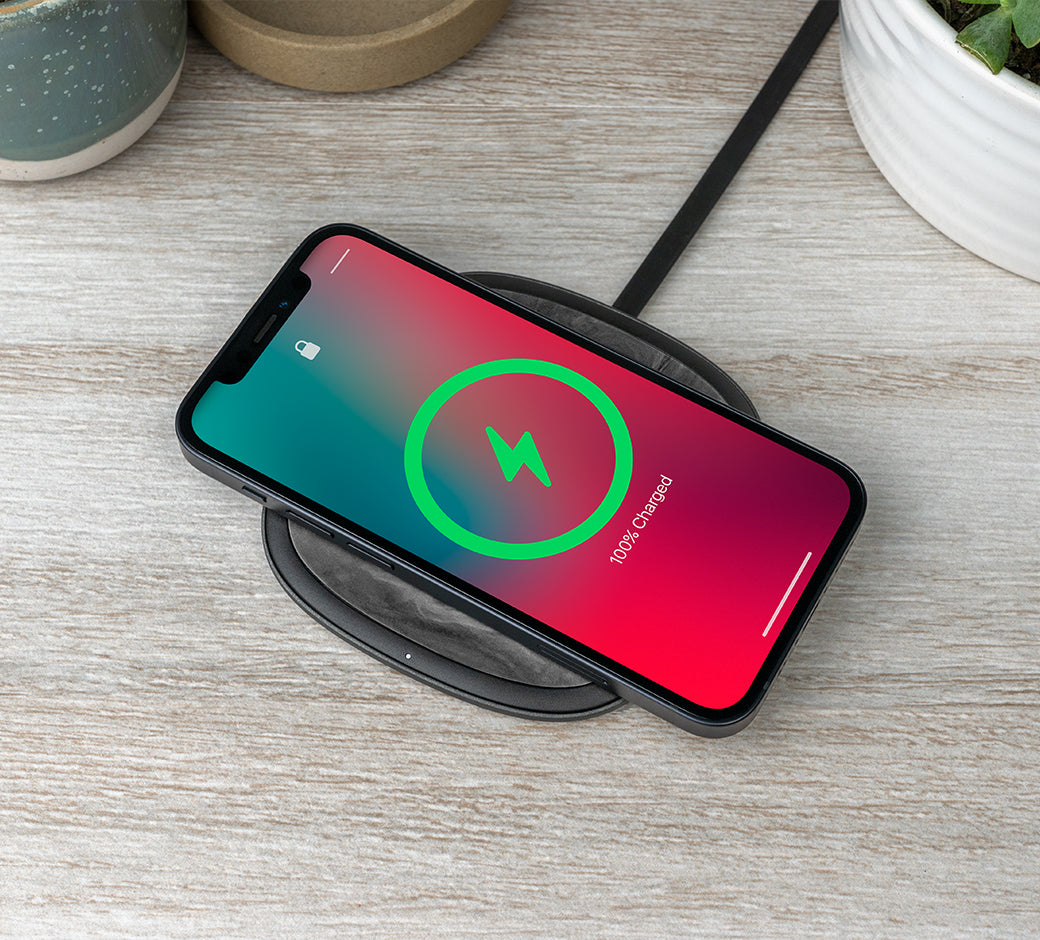 A smartphone wirelessly charging on a black charging pad placed on a light wood surface, surrounded by decorative plants.