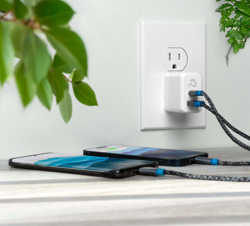 White wall charger with dual cables charging two smartphones on a desk with green plants nearby.
