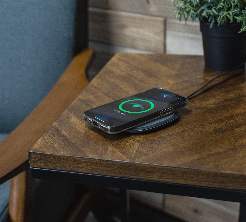 A smartphone wirelessly charging on a sleek black pad, placed on a wooden table near a potted plant and chair.