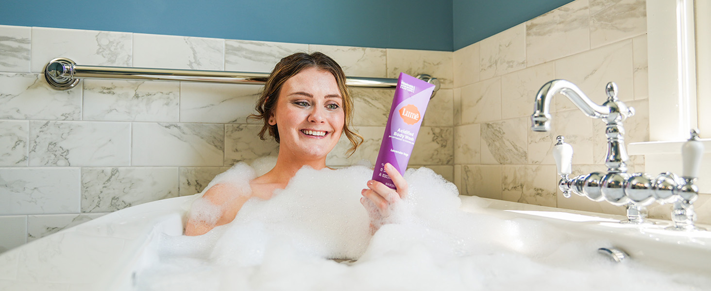 A woman using Lumē Acidified Body Wash in Lavender Sage being used in a bath tub