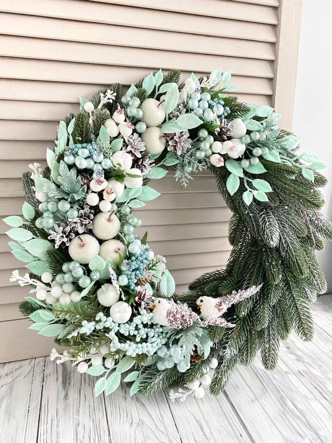 May include: A green and white Christmas wreath with white berries, pine cones, and small white apples. Two white birds are perched on the bottom of the wreath.